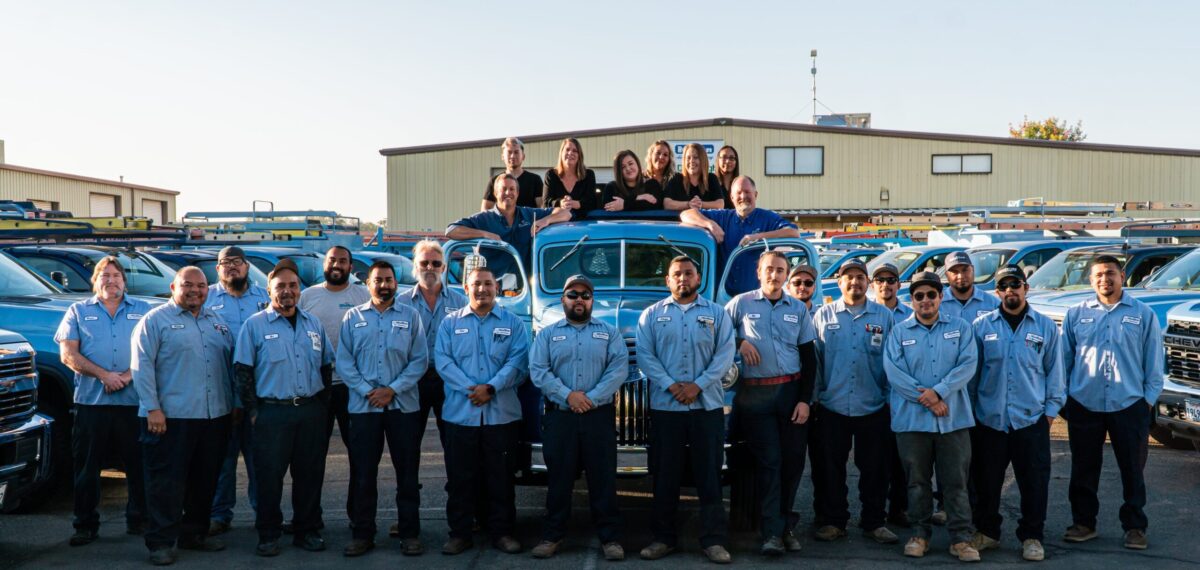 A large group portrait of the JC Melton AC repair team, featuring many technicians in blue uniforms standing in front of a row of blue service trucks and an antique blue pickup truck, with several non-uniformed office staff standing or leaning on the antique truck