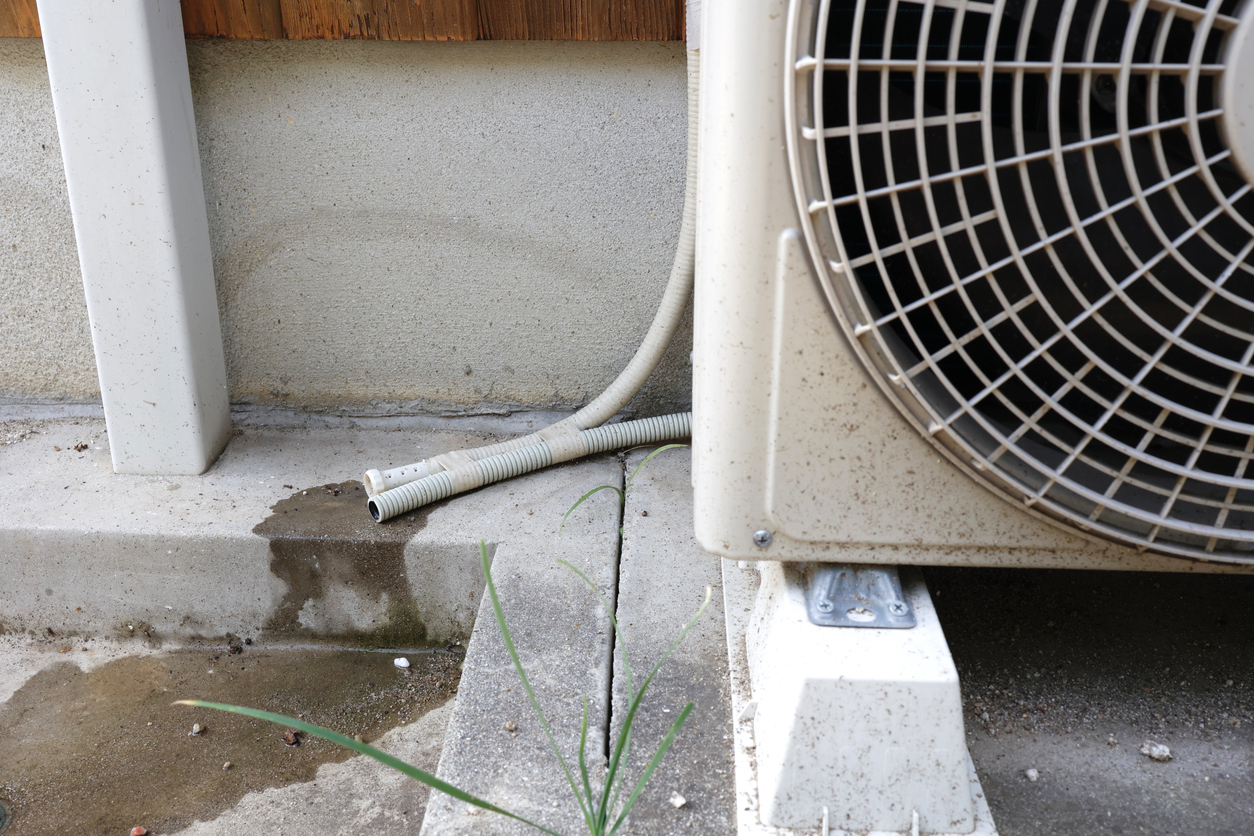 A close-up, ground-level image showing the exterior condensate drain lines of an AC unit dripping water onto the concrete pad below, indicating a need for AC repair or maintenance from JC Melton.