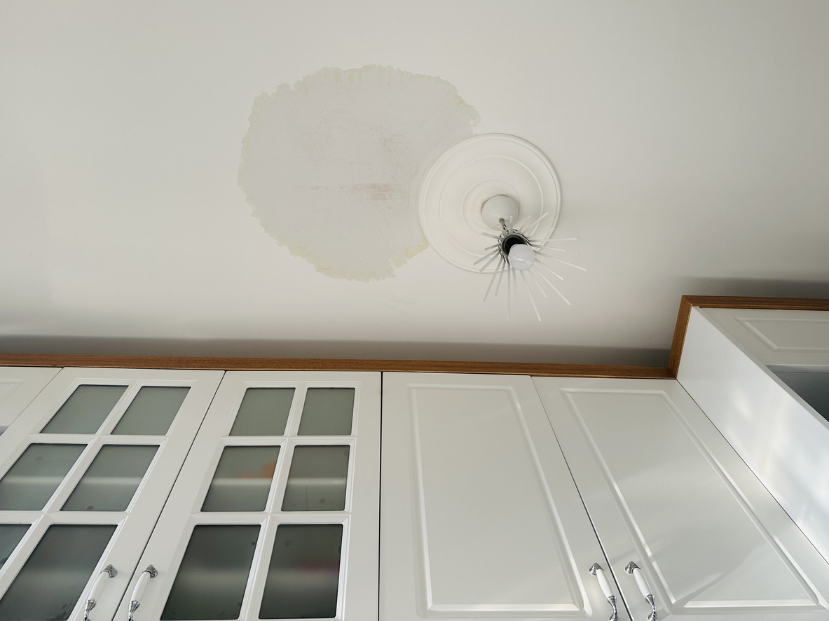 An upward-angle shot of a white kitchen ceiling showing a large, circular brown water stain around a decorative light fixture, signaling a potential AC leak issue requiring JC Melton