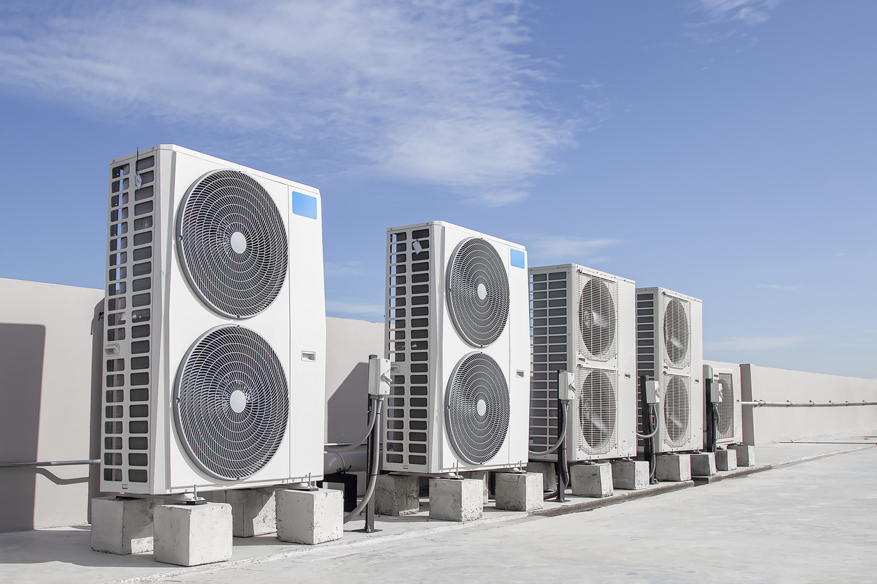 A line of multiple large commercial or multi-unit AC condensing units is installed on a flat rooftop on concrete blocks against a clear blue sky, demonstrating the commercial HVAC services offered by JC Melton.