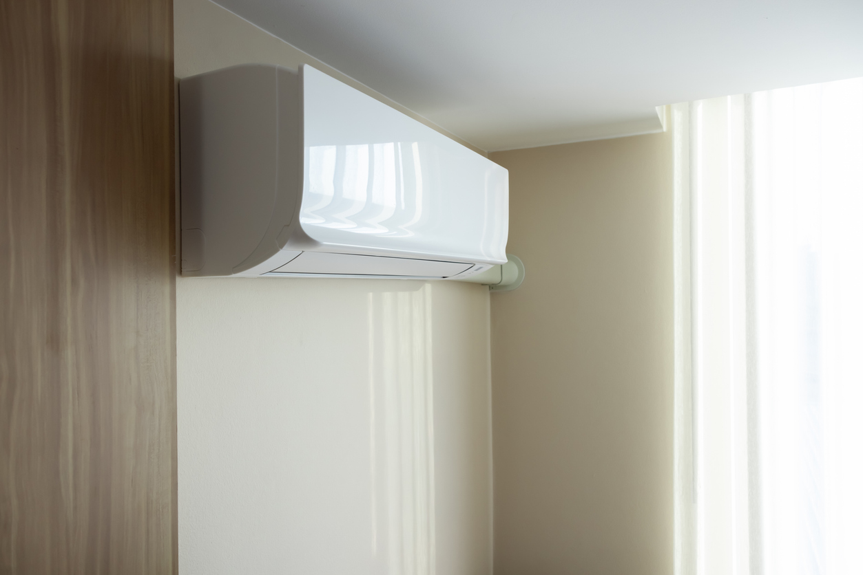 A white, wall-mounted mini-split air conditioner indoor unit is installed in the corner of a room with light beige walls and a light wood paneling, highlighting a type of HVAC system that JC Melton is equipped to repair and maintain.