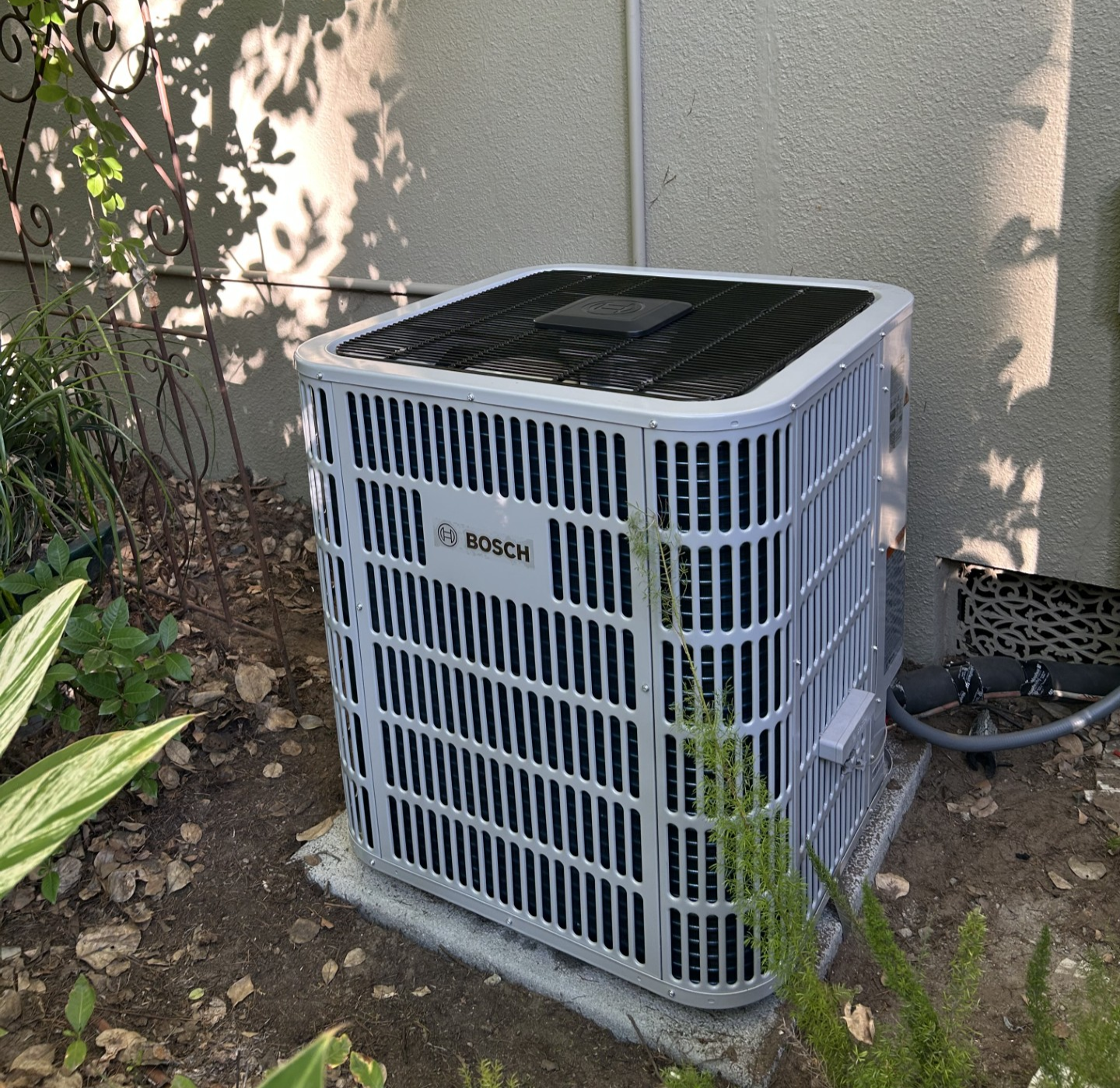 A white, square Bosch split-system HVAC outdoor condenser unit sitting on a concrete pad next to a light-colored building wall, surrounded by dirt and landscaping plants.