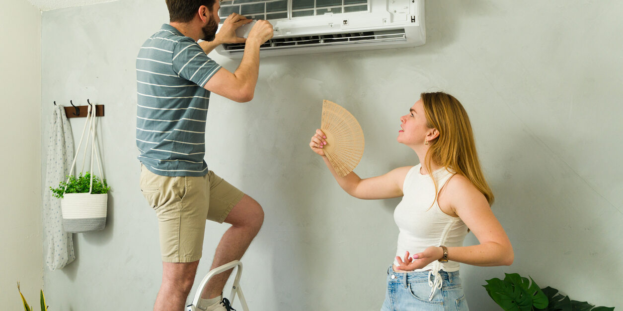young couple fixing an air conditioning unit while woman holds hand fan